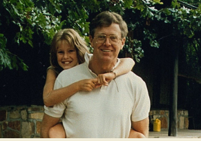 Charles Koch wearing a white polo and glasses carrying daughter Elizabeth on his back, both smiling, with trees in the background.